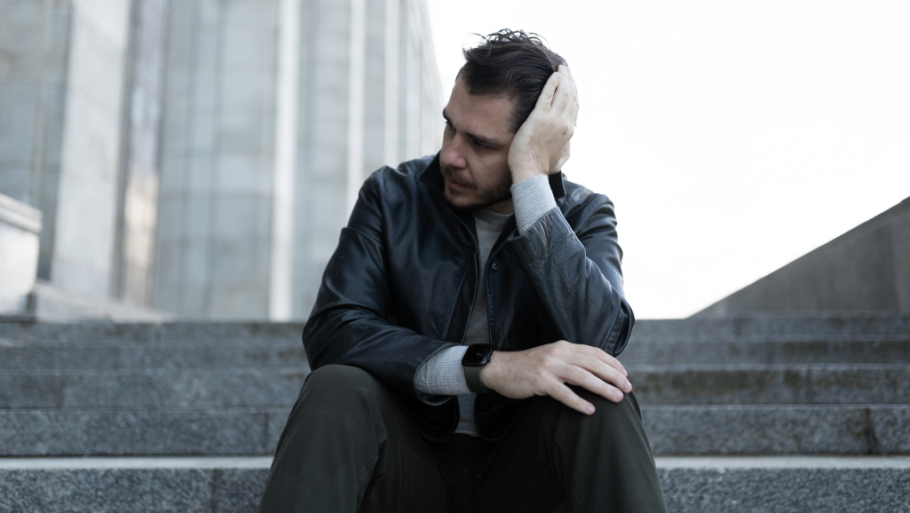 A detailed full-body portrait of a person experiencing physical symptoms of stress. The subject is standing against a plain grey background, lit from the side to create dramatic shadows and highlights. Their face is furrowed, brow creased, lips pursed in tension. Veins are visibly pulsing in their temples and neck. Their shoulders are hunched, hands clenched into tight fists. The pose conveys a sense of anxiety, with the subject's body language reflecting the internal strain of high stress levels. The image has a somber, melancholic mood, reflecting the negative physical impacts of prolonged stress.