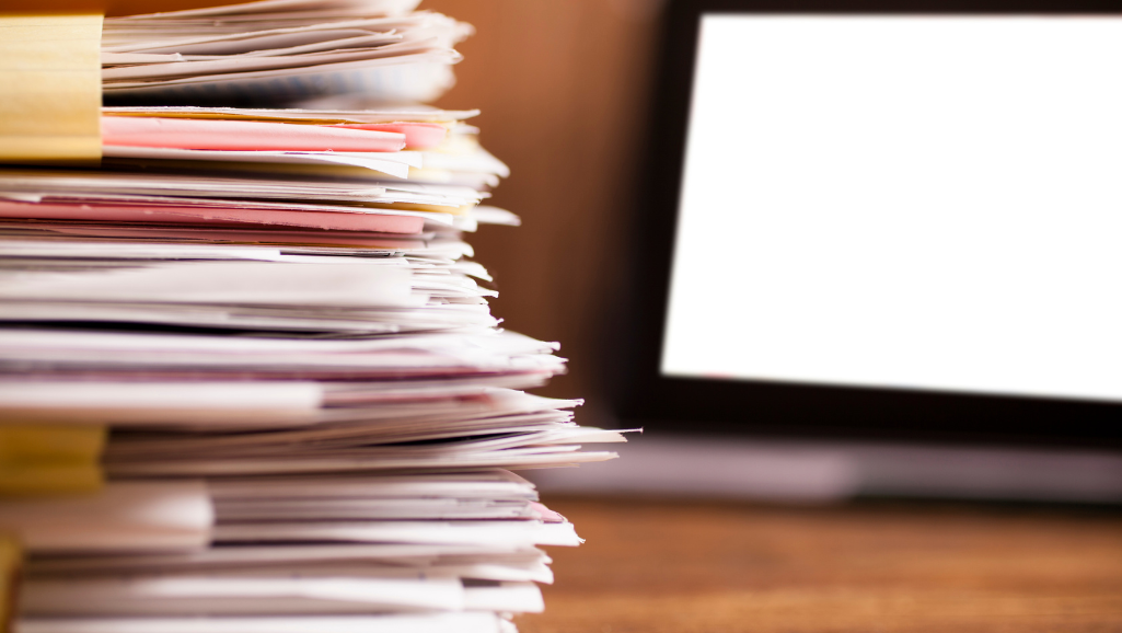 A dimly lit office space, cluttered with overflowing paperwork, stacks of books, and a tangle of cables. In the foreground, a desk lamp casts a warm glow, illuminating a computer screen displaying multiple open windows and notifications. The middle ground features a desk overflowing with sticky notes, a spilled cup of coffee, and a phone ringing incessantly. In the background, the walls are adorned with framed certificates and awards, partially obscured by the chaos. The overall atmosphere conveys a sense of overwhelming stress and pressure, with a muted color palette and heavy shadows contributing to the anxious mood.