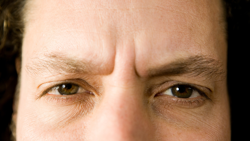 A close-up view of a person's face, eyes strained and brow furrowed, conveying a sense of mental exhaustion and distress. The skin appears taut, with visible signs of fatigue and worry lines. The lighting is low and moody, casting dramatic shadows that accentuate the tension in the subject's expression. The background is blurred, hazy, and indistinct, placing the focus entirely on the central figure's face and the visible symptoms of chronic stress.